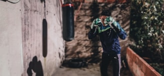 A person wearing a dark jacket and green gloves is standing in a fighting stance in front of a punching bag. The environment appears to be an outdoor area with a textured wall and some greenery in the background. Shadows are cast on the wall from a light source to the left. The mood is focused and dynamic.