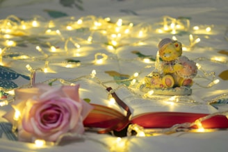 A cozy scene featuring an open book surrounded by warm, glowing fairy lights. A cute teddy bear figurine is seated on the pages, adding a whimsical touch. A pink rose rests nearby, enhancing the soft and calming atmosphere.