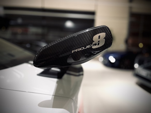 A close-up view of a car's side mirror with 'Project 8' written on it. The mirror has a carbon fiber texture and is attached to a sleek white car, with a blurred showroom background.