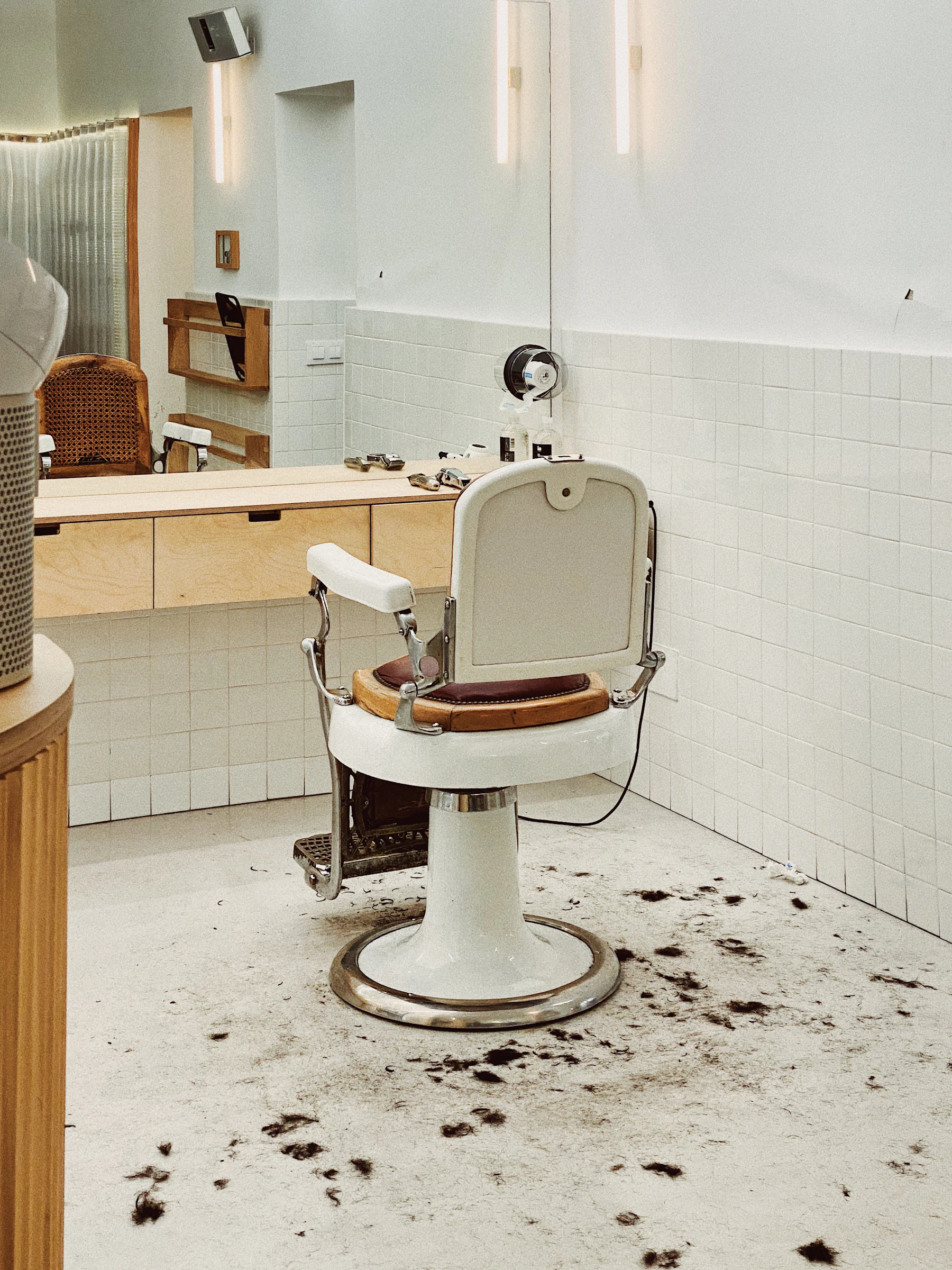 A vintage cream barber's chair in a white-tiled room, hair on the floor around it