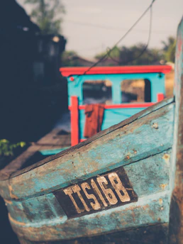 A rustic wooden boat painted in turquoise and red, with a prominent identification number 'TTS16B' on its side. The background is blurred, suggesting a setting near water with hints of natural greenery and possibly structures.