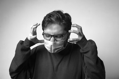 A person wearing glasses and a medical mask stands with their eyes closed and hands raised to their head, conveying a sense of tension or frustration. The person is dressed in a dark sweater and is positioned against a plain, light-colored background in a black and white photograph.