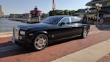 A luxurious black car is parked on a brick-paved waterfront street. In the background, there is a red lighthouse, several buildings, and a restaurant. The car has shiny chrome accents and is oriented towards the right, under a clear, sunny sky.