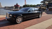 A luxurious black car is parked on a brick-paved waterfront street. In the background, there is a red lighthouse, several buildings, and a restaurant. The car has shiny chrome accents and is oriented towards the right, under a clear, sunny sky.