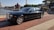 A luxurious black car is parked on a brick-paved waterfront street. In the background, there is a red lighthouse, several buildings, and a restaurant. The car has shiny chrome accents and is oriented towards the right, under a clear, sunny sky.