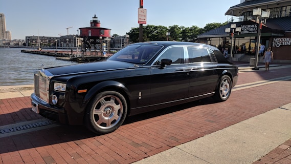 A luxurious black car is parked on a brick-paved waterfront street. In the background, there is a red lighthouse, several buildings, and a restaurant. The car has shiny chrome accents and is oriented towards the right, under a clear, sunny sky.