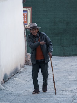 An elderly person is walking along a narrow street, using a wooden cane for support. The individual is wearing a gray fedora hat, a dark coat over an orange sweater, and loose-fitting pants. The background shows a white wall and a partially visible sign with some text.