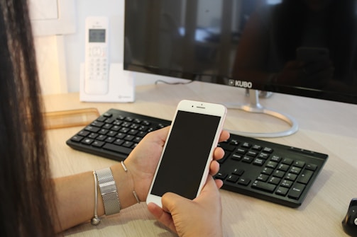 A person is holding a smartphone while sitting at a desk with a keyboard, mouse, and a monitor displaying the brand 'KUBO'. A cordless phone is also visible in the background.