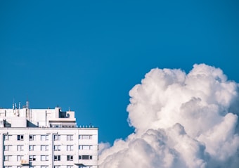 A modern white apartment building with multiple windows, located on the left side of the image under a clear blue sky. To the right, large, fluffy white clouds fill the scene, appearing vibrant and dense.