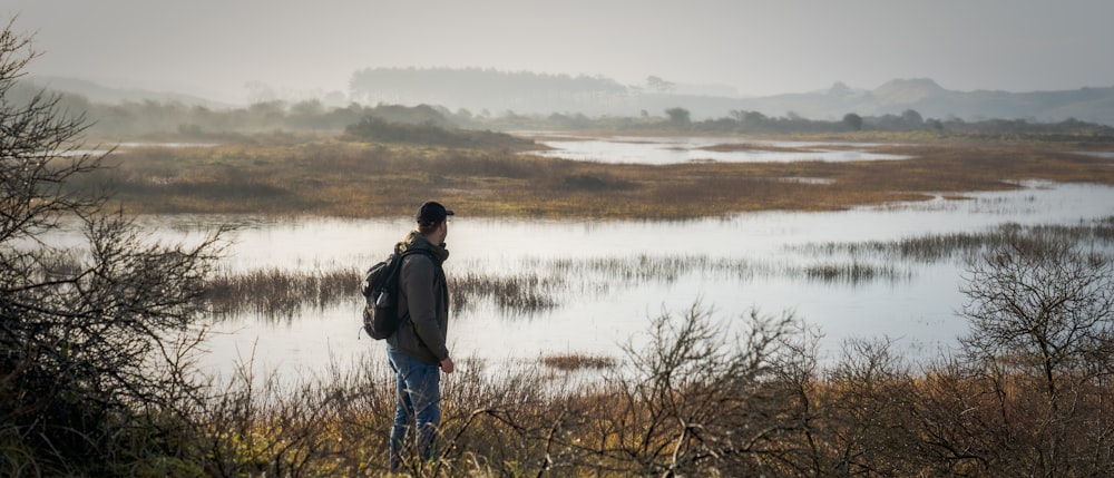Uomo in giacca nera in piedi sull'erba marrone vicino al lago durante il giorno
