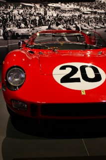 A vintage red racing car with a large white circle and the number 20 painted on the hood. The car is displayed in a museum setting, with a black and white photograph of a racing event in the background featuring a large crowd.