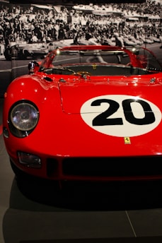 A vintage red racing car with a large white circle and the number 20 painted on the hood. The car is displayed in a museum setting, with a black and white photograph of a racing event in the background featuring a large crowd.