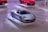 A sleek silver sports car is prominently displayed on a white platform in a showroom. The background features another red sports car on a similar platform. The floor has yellow and orange circular and spiral patterns.