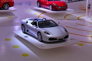 A sleek silver sports car is prominently displayed on a white platform in a showroom. The background features another red sports car on a similar platform. The floor has yellow and orange circular and spiral patterns.