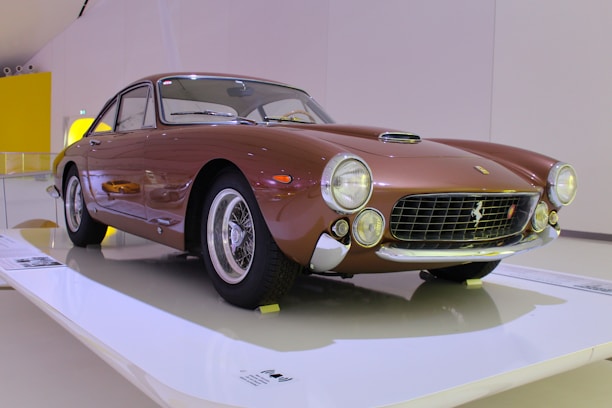 A classic brown sports car displayed in a well-lit showroom. The car features round headlights, a sleek aerodynamic design, and polished chrome elements. It is elevated on a platform with information panels nearby.