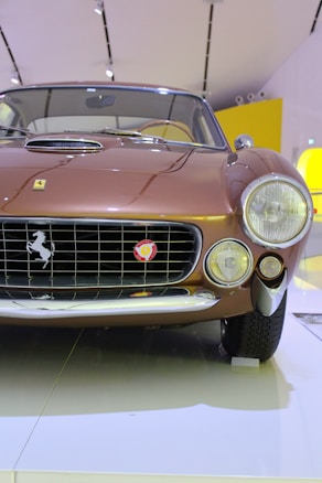 Front view of a classic Ferrari car with a brown metallic finish, showcasing its iconic grille and emblem, large circular headlights, and polished chrome details. The background appears to be an indoor setting with white and yellow elements visible.
