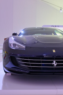 A sleek, black luxury sports car is displayed. The car features a prominent emblem on the front, smooth aerodynamic lines, and a shiny exterior. It is positioned indoors under bright lighting, highlighting its glossy finish and modern design.