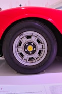 A close-up view of a car wheel with a modern alloy rim design, featuring a yellow center cap with stylized branding. The wheel is set against a vibrant red car body, which curves elegantly over the tire. The background is blurred with text partially visible on a surface below the wheel.