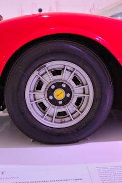 A close-up view of a car wheel with a modern alloy rim design, featuring a yellow center cap with stylized branding. The wheel is set against a vibrant red car body, which curves elegantly over the tire. The background is blurred with text partially visible on a surface below the wheel.