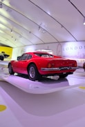 A vintage red sports car is showcased in a modern, bright exhibition space with a sleek, curved ceiling. The wall displays landmarks and city names, including Paris and London, suggesting a thematic or historical presentation.