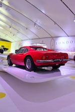 A vintage red sports car is showcased in a modern, bright exhibition space with a sleek, curved ceiling. The wall displays landmarks and city names, including Paris and London, suggesting a thematic or historical presentation.