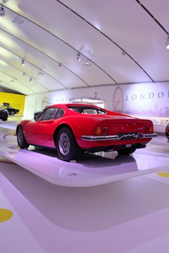 A vintage red sports car is showcased in a modern, bright exhibition space with a sleek, curved ceiling. The wall displays landmarks and city names, including Paris and London, suggesting a thematic or historical presentation.