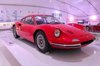 A classic red sports car is displayed in a modern, well-lit showroom. The car's sleek design and polished surface highlight its vintage elegance. In the background, several posters and cars are visible, adding to the exhibition ambiance.