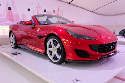 A sleek red sports car with a convertible design is displayed in a modern showroom. The car features elegant curves, prominent air intakes, and a distinctive badge on the hood, emphasizing its luxury and performance-oriented nature. The setting is well-lit with a minimalistic background, highlighting the vehicle.