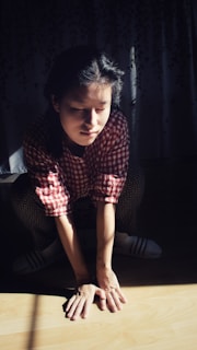 A person in a red and white checkered shirt squats on a wooden floor with their hands resting on the ground. Sunlight creates a high contrast, illuminating parts of their face and arms while casting shadows in the background.