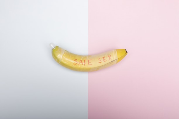 A banana with the words 'SAFE SEX' written on it is wrapped with a condom. The background is split into two halves, one white and the other pink.