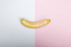 A banana with the words 'SAFE SEX' written on it is wrapped with a condom. The background is split into two halves, one white and the other pink.