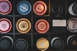 A grid of various bottle caps arranged in a square black box, each cap with unique designs and colors including red, gold, blue, and black. The caps have intricate logos and branding, suggesting they are from different beverage brands.