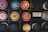 A grid of various bottle caps arranged in a square black box, each cap with unique designs and colors including red, gold, blue, and black. The caps have intricate logos and branding, suggesting they are from different beverage brands.