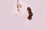 A bear plushie is looking at a doll suspended by black straps against a light pink background. The doll has blonde hair and is posed in a seated position with its arms and legs restrained.