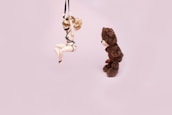 A bear plushie is looking at a doll suspended by black straps against a light pink background. The doll has blonde hair and is posed in a seated position with its arms and legs restrained.