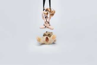 A small, articulated doll with blonde hair is suspended by black straps, positioned above a teddy bear that is resting on the ground. The teddy bear is light brown and fluffy, wearing a striped bow tie. The background is a plain, pale blue.