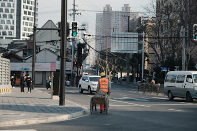 An urban street scene with a construction worker pushing a cart wearing an orange safety vest and hard hat. The streets are lined with modern and traditional buildings. There are cars and a van on the road, and pedestrians on the sidewalks. A traffic light is visible, showing green. Leafless trees suggest a winter setting.