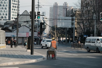 An urban street scene with a construction worker pushing a cart wearing an orange safety vest and hard hat. The streets are lined with modern and traditional buildings. There are cars and a van on the road, and pedestrians on the sidewalks. A traffic light is visible, showing green. Leafless trees suggest a winter setting.
