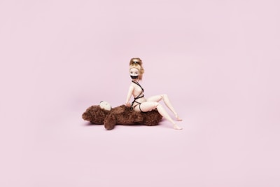 A doll wearing a black bondage-themed outfit is posed sitting on a brown stuffed teddy bear against a soft pink background. The doll is styled with light-colored hair tied up.