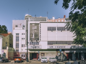 A vintage-style cinema building with a white facade displays film posters and a marquee sign with the text 'UM IVANOV.' Several cars are parked in front of the cinema, and there are trees on both the sides. The sky is clear, and it appears to be a sunny day.