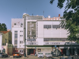 A vintage-style cinema building with a white facade displays film posters and a marquee sign with the text 'UM IVANOV.' Several cars are parked in front of the cinema, and there are trees on both the sides. The sky is clear, and it appears to be a sunny day.