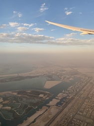 Aerial view capturing a coastal cityscape with intricate land formations and water bodies. The wing of an airplane is visible in the upper right, set against a backdrop of a partly cloudy sky. The horizon is hazy, blending smoothly with the earth tones below.