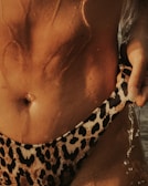 A close-up of a person's midsection featuring wet skin glistening in the light. A gold belly ring is visible. The person is wearing leopard print swimwear, and water droplets are cascading down their body.