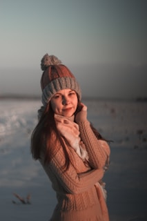 A person wearing a knitted hat with a pom-pom and a warm sweater stands outdoors with a snowy background. The lighting suggests a sunset or sunrise, casting a warm glow on the individual's face and surrounding.