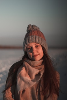 A person is dressed warmly in winter attire, including a knitted hat with a pom-pom and a cozy scarf, set against a snowy outdoor backdrop. Long hair flows down over the warm clothing.