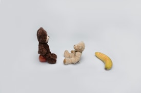 Two teddy bears sit facing each other on a light grey background. The dark brown bear is seated on a red object. Nearby, a banana with the words 'SAFE SEX' written on it lies on the surface.