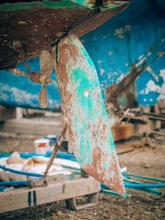 A close-up view of the underside of a boat reveals a worn and rusted metal rudder and propeller. The boat's hull has chipped and peeling blue and green paint, suggesting age and exposure to the elements. The scene is set in a shipyard or repair area, with tools and materials scattered around.