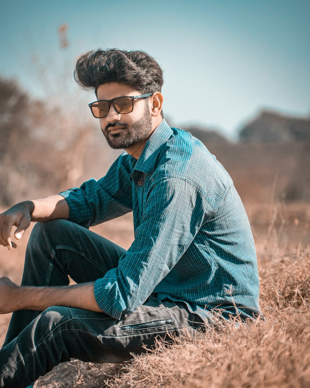 man in blue dress shirt and blue denim jeans sitting on brown grass during daytime