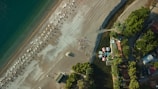 An aerial view of a beach with a long stretch of shoreline, featuring neatly arranged rows of sunbeds and umbrellas. People are gathered in a small formation near the water's edge. The sea appears calm with a gradient of blue and green. Towards the right, there are green trees and a cluster of buildings with colorful roofs lining the beach.