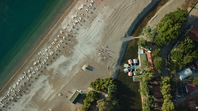 An aerial view of a beach with a long stretch of shoreline, featuring neatly arranged rows of sunbeds and umbrellas. People are gathered in a small formation near the water's edge. The sea appears calm with a gradient of blue and green. Towards the right, there are green trees and a cluster of buildings with colorful roofs lining the beach.